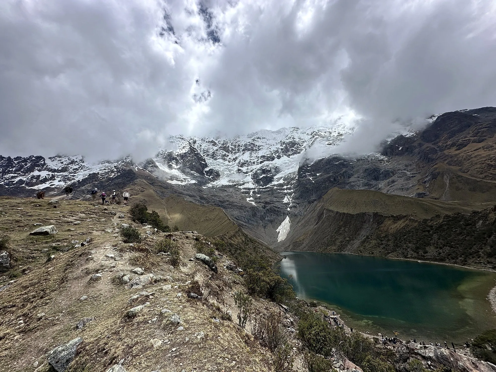 Walkers on the ridge above the turquoise Humantay glacial lake, under snow peaks in cloud near Cusco