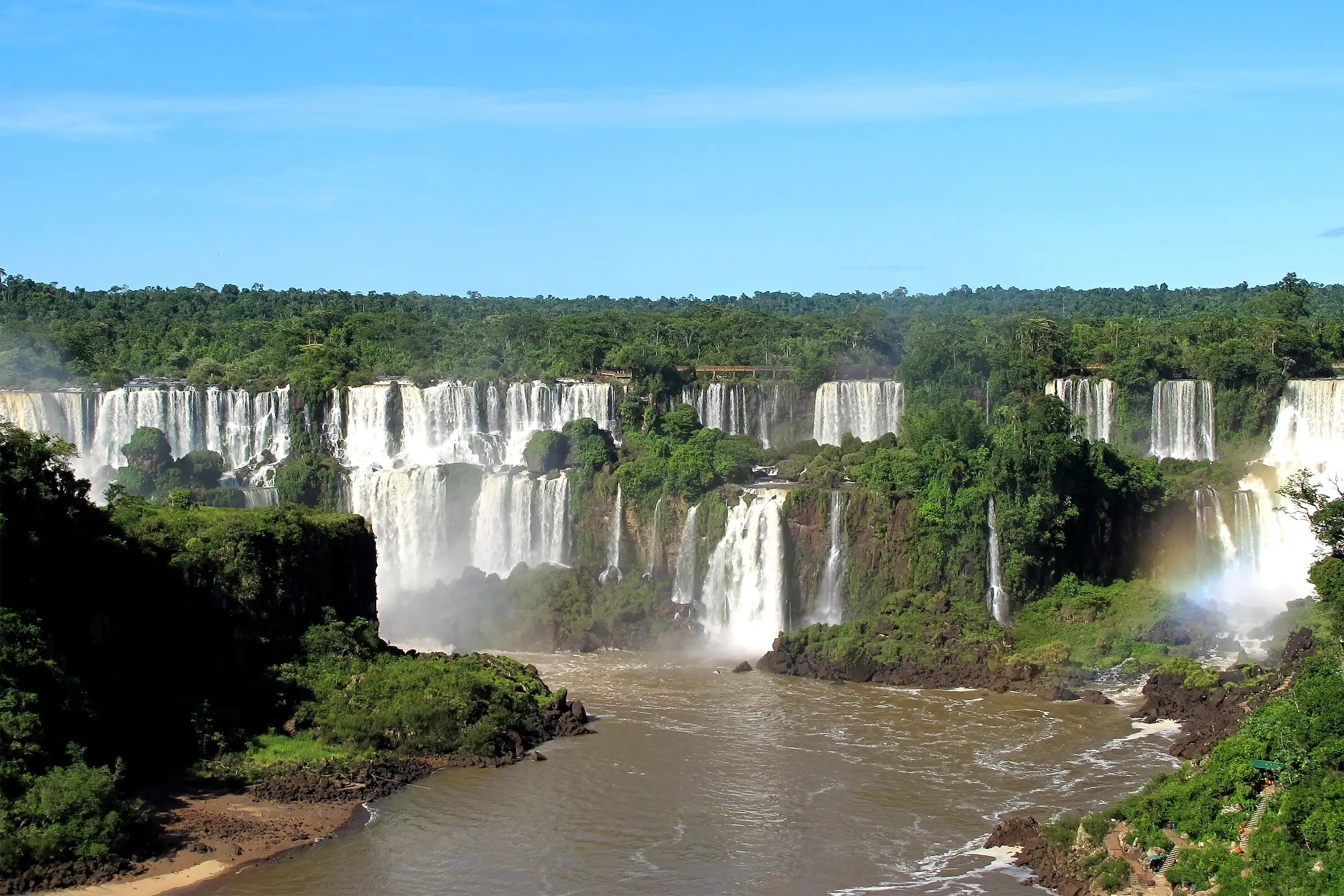 The Iguazu falls seen from the Brazilian side, a wall of waterfalls in the forest above a brown river with a rainbow on the right