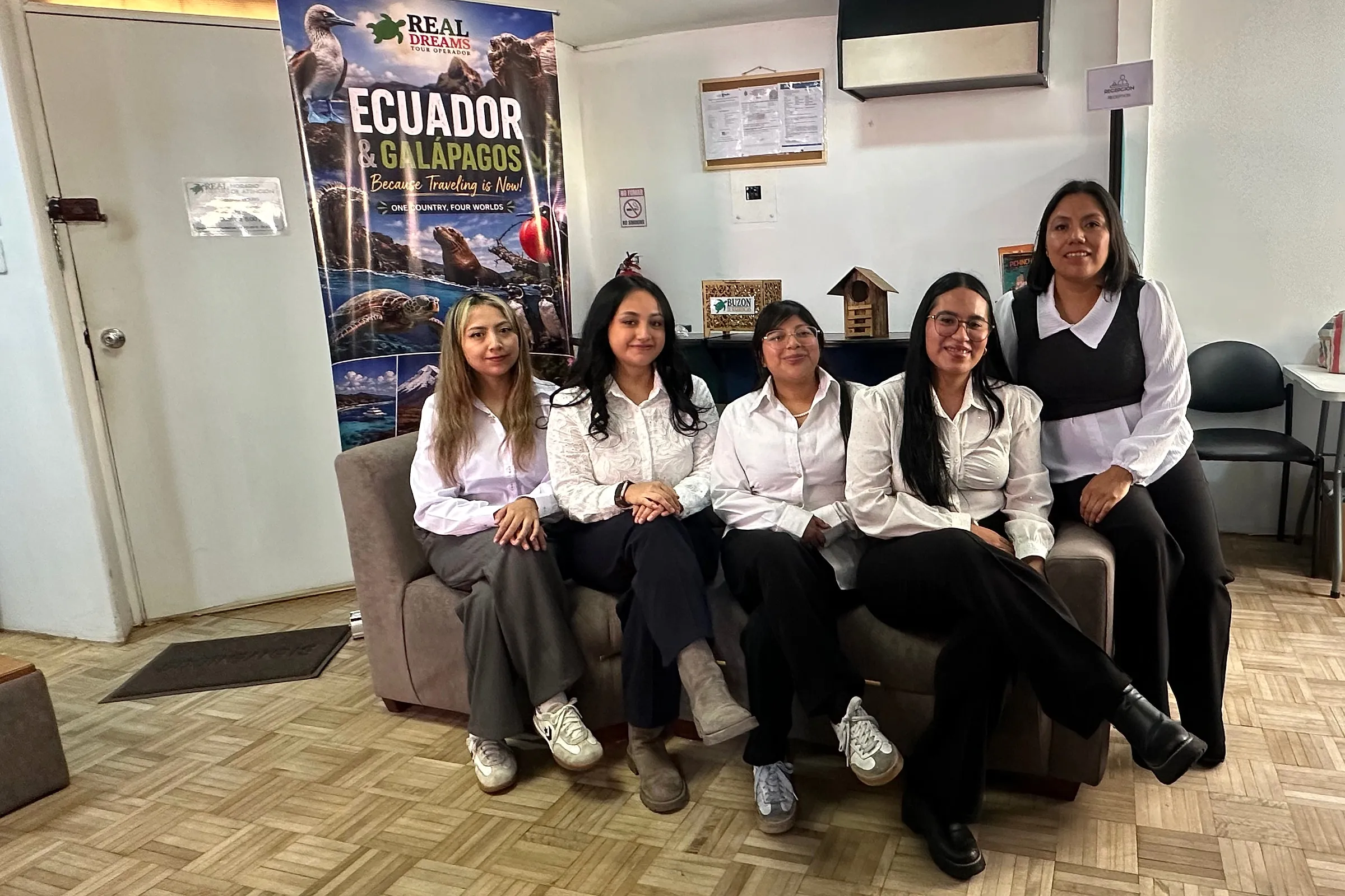 Five members of the Latin Lands by Real Dreams team sitting on a sofa beside an Ecuador and Galápagos banner at the office in Quito