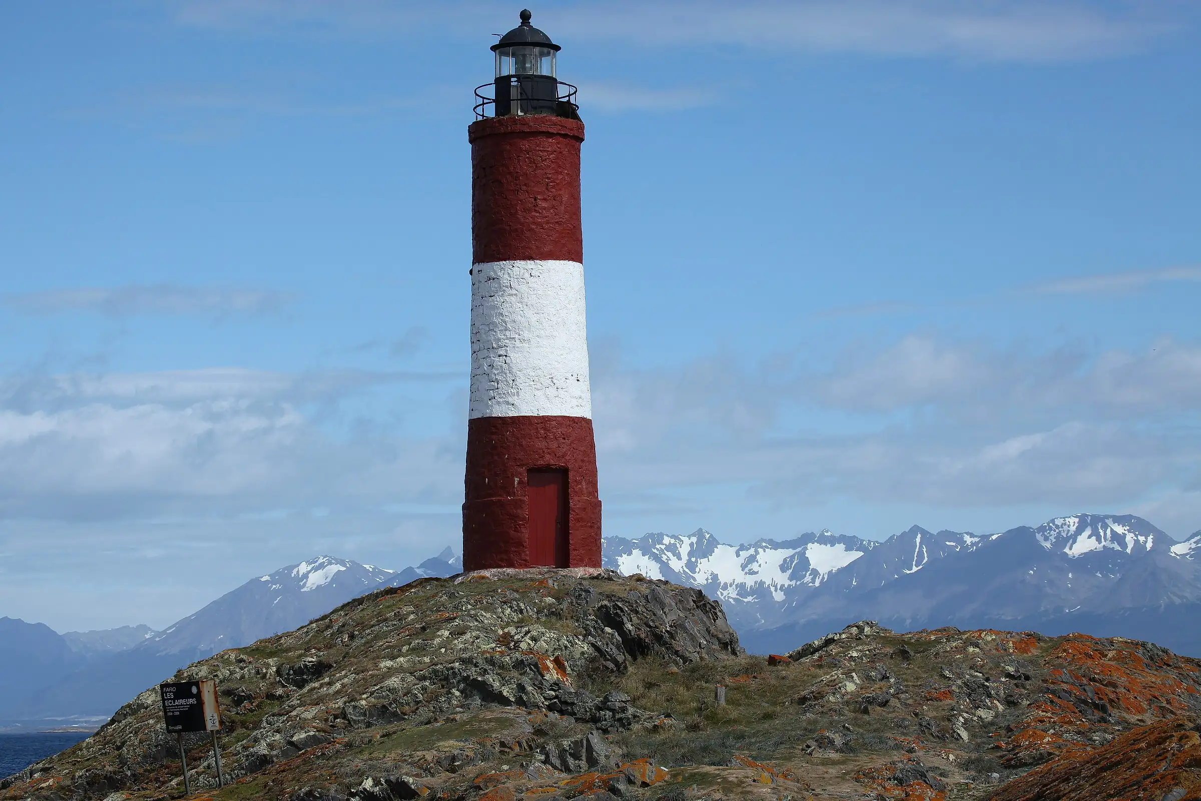 The red and white Les Eclaireurs lighthouse on a rocky islet in the Beagle Channel, with snowy peaks behind, near Ushuaia