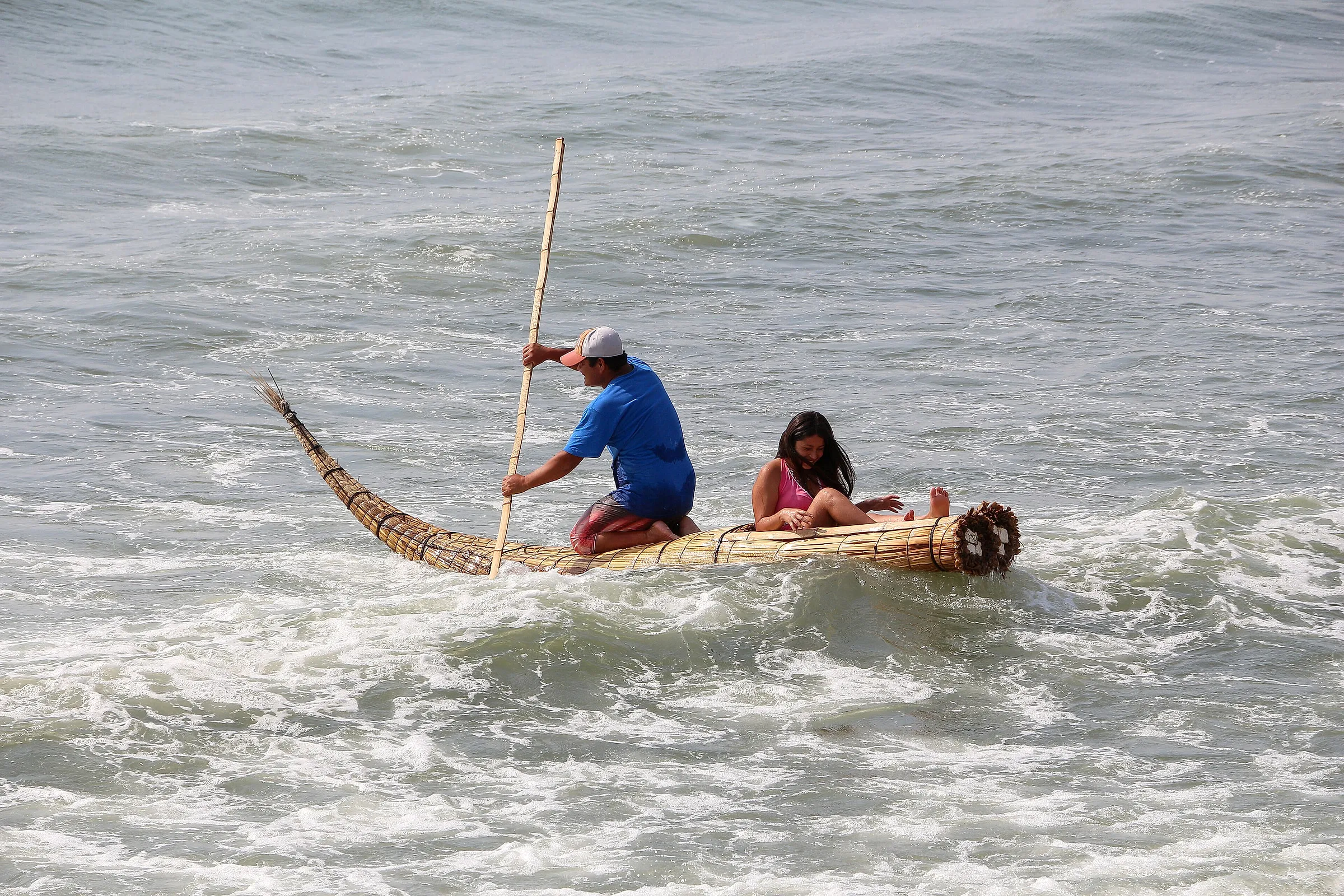 A fisherman paddling a caballito de totora reed boat through the surf at Huanchaco, with a passenger