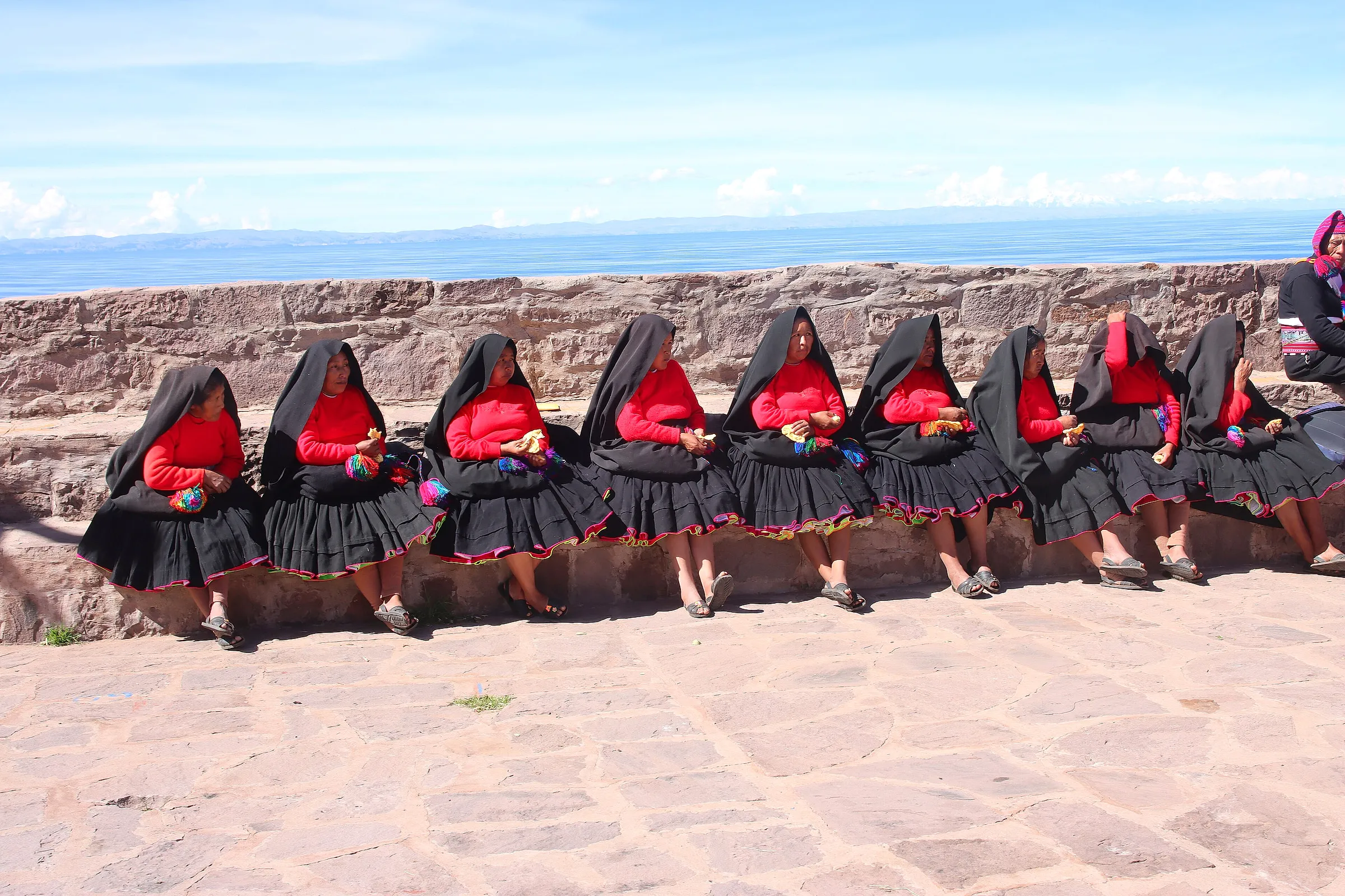 Women from Taquile island in traditional black and red dress, seated on a stone wall above Lake Titicaca
